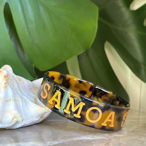 Könnte beinhalten: Ein Armband mit Schildpattmuster und dem Wort "SAMOA" in goldfarbenen Buchstaben. Das Armband befindet sich vor einem Hintergrund aus grünen Blättern und einer Muschel, was eine tropische Ästhetik erzeugt.