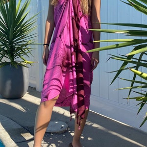 Op de afbeelding: Een roze en maroon sarong met een grote bloemenprint. De sarong is om de taille gebonden en heeft een franje aan de onderkant.