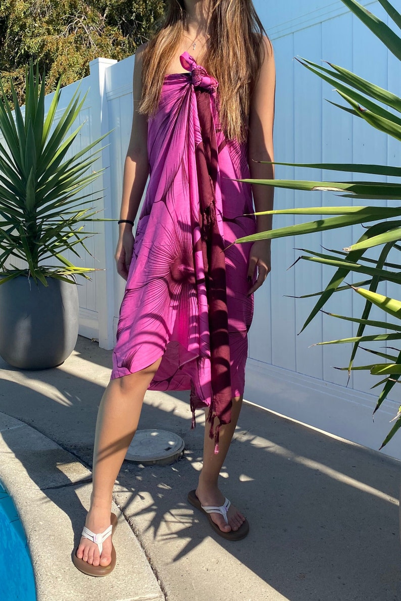 Op de afbeelding: Een roze en maroon sarong met een grote bloemenprint. De sarong is om de taille gebonden en heeft een franje aan de onderkant.