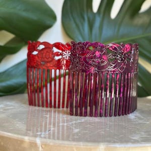 Puede incluir: Dos peines decorativos para el cabello. Uno es rojo con un diseño floral, y el otro es magenta oscuro con un patrón floral similar. Los peines están hechos de un material translúcido y se encuentran sobre una superficie similar al mármol.