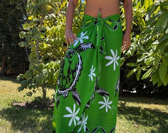 Op de afbeelding: Een groene sarong met witte bloemen en zwarte schildpadmotieven. De sarong is om de taille geknoopt en wordt gedragen over een zwarte tube top. De stof is felgroen, met witte bloemen en zwarte schildpadmotieven.