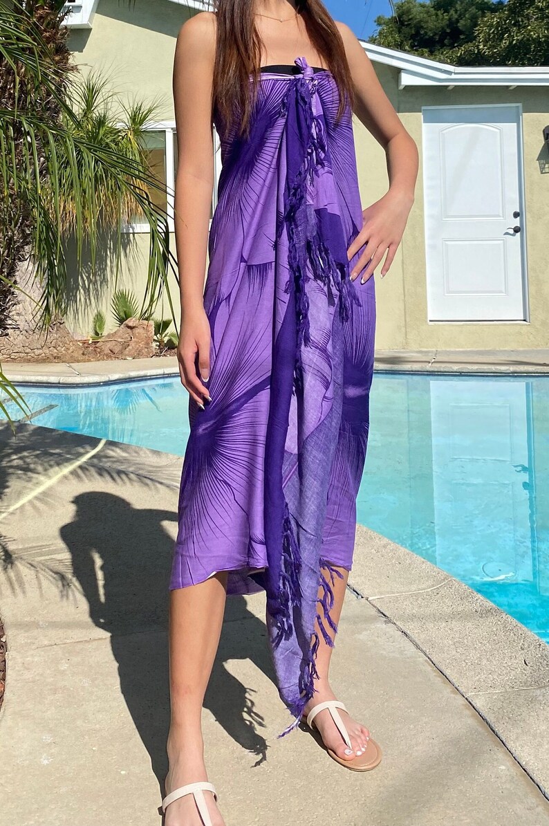 Op de afbeelding: Een paarse sarong met een bloemenprint en franjes. De sarong is in de taille geknoopt en wordt als jurk gedragen.