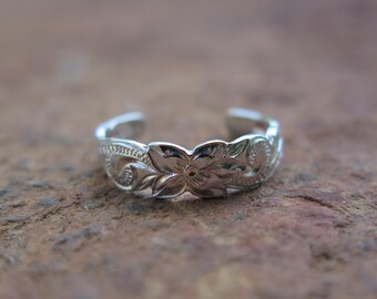 May include: A silver-colored toe ring with a floral design. The ring features an open design and is adorned with detailed carvings of flowers and leaves. The ring is displayed on a brown surface.