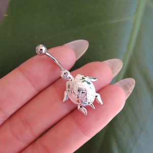 May include: A silver-colored belly button ring featuring a turtle design. The turtle has a textured shell and small flipper details. The curved barbell has a small ball at the top.