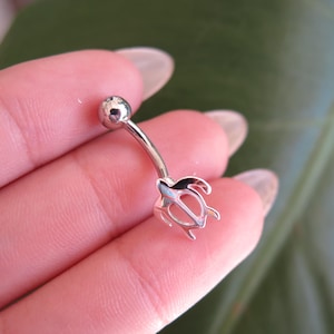 May include: A silver-colored belly button ring featuring a turtle design. The curved barbell has a small ball on one end and a turtle-shaped charm on the other. The turtle charm has a detailed, openwork design.