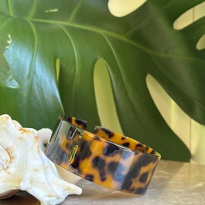 Può includere: Un bracciale a polsino con motivo tartarugato e finitura lucida. Il bracciale è aperto nella parte inferiore e poggia su una superficie chiara accanto a una conchiglia. Una grande foglia verde è sullo sfondo.