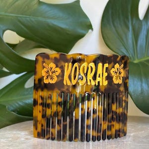 Puede incluir: Un peine para el cabello con estampado de carey con la palabra "KOSRAE" en letras doradas. Dos diseños de flores de hibisco doradas flanquean el texto. El peine tiene una parte superior rectangular y dientes verticales.