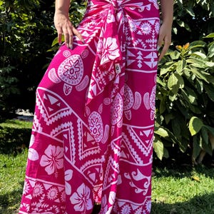 May include: A vibrant fuchsia sarong featuring a white graphic print of sea turtles, hibiscus flowers, and geometric tribal patterns. The fabric is tied at the waist with a central knot, creating a draped, floor-length silhouette with wide legs.