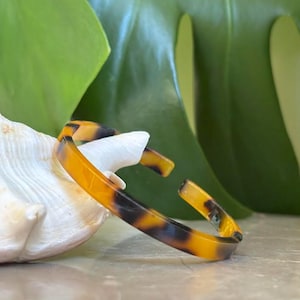 May include: A tortoiseshell-patterned bangle bracelet with an open design. The bracelet features shades of brown and amber, with a glossy finish. The jewelry is displayed near a seashell and green foliage.