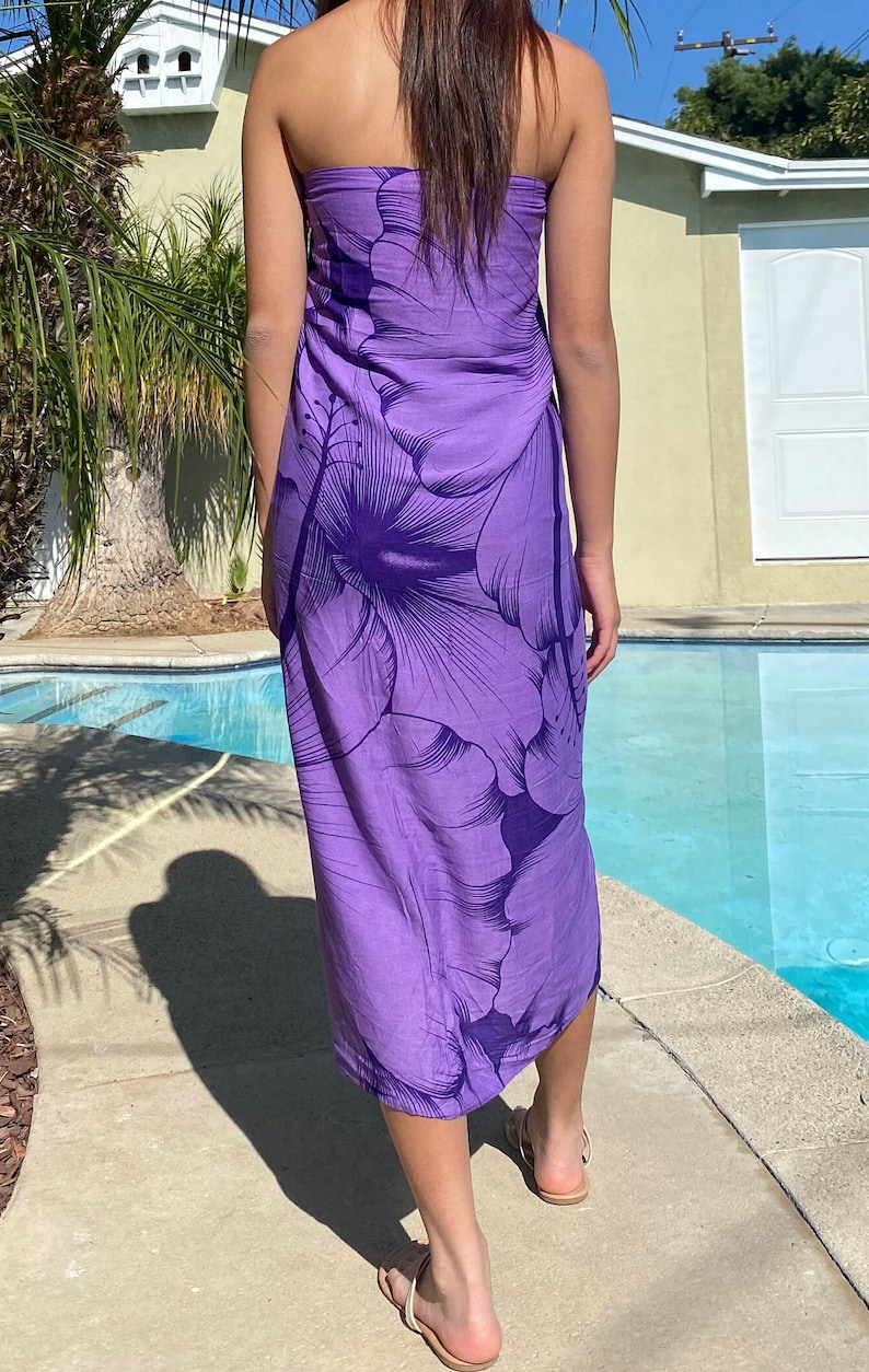 Op de afbeelding: Een paarse sarong met een groot hibiscusbloemmotief. De sarong is om de taille gebonden en valt tot aan de enkels.