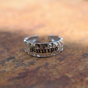 May include: A silver-colored ring with the word "Ku'uipo" engraved in black, stylized lettering. The ring has a textured surface and an open design. The ring is displayed on a brown surface.