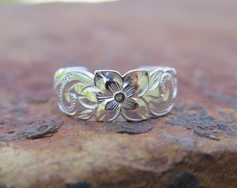 May include: A silver-colored ring with a floral design. The ring features a central flower with detailed petals and surrounding embossed patterns. The ring is set against a textured, brown surface.