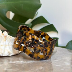 May include: A wide, tortoise-shell patterned bracelet with a floral and turtle design. The bracelet is made of a glossy, translucent material. The bracelet is approximately 2 inches wide.