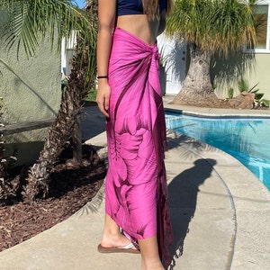 Op de afbeelding: Een roze sarong met een donker bloemenprint. De sarong is om de taille gebonden en heeft een franje aan de onderkant.