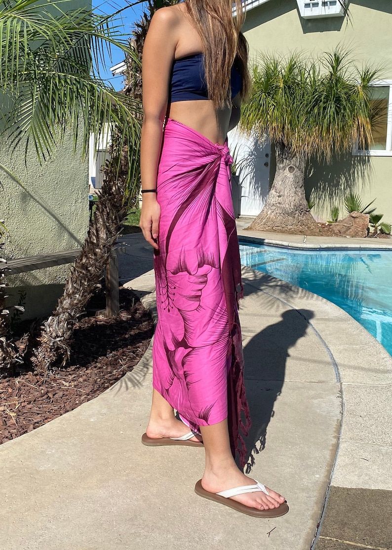 Op de afbeelding: Een roze sarong met een donker bloemenprint. De sarong is om de taille gebonden en heeft een franje aan de onderkant.