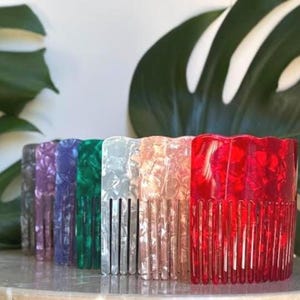 Puede incluir: Una fila de peines para el cabello coloridos en tonos morado, azul, verde, blanco, rosa y rojo. Cada peine tiene un borde superior ondulado y dientes verticales. Los peines están hechos de un material translúcido y veteado.