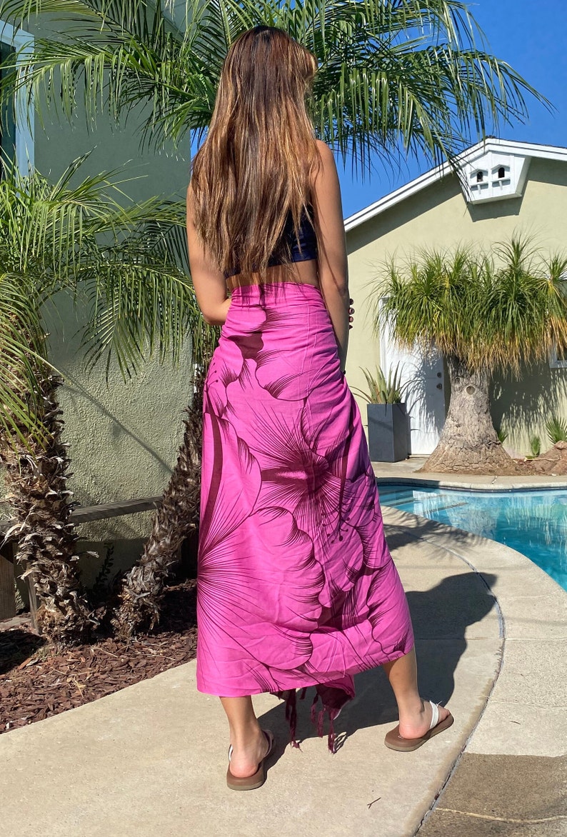 Op de afbeelding: Een roze sarong met een grote donkerbruine bloemenprint. De sarong is om de taille gebonden en heeft een franje aan de onderkant.