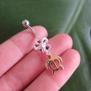 May include: A silver belly button ring with a flower and a gold-colored turtle charm. The flower has pink and silver petals. The jewelry is designed for body piercing and is shown against a green background.