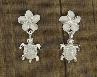 May include: A pair of silver earrings. Each earring has a flower-shaped top with a small gem, connected to a dangling turtle charm. The turtle charms have a textured surface, creating a detailed design.