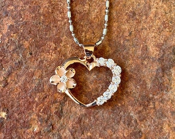 Puede incluir: Un collar con colgante de corazón dorado con una flor y cristales transparentes. El corazón es abierto, con cristales a un lado. La flor tiene un centro y pétalos de color rosa claro. La cadena del collar es plateada.