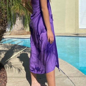 Op de afbeelding: Een paarse sarong met een bloemenprint. De sarong is om de taille gebonden en valt tot aan de enkels. De persoon draagt witte sandalen.
