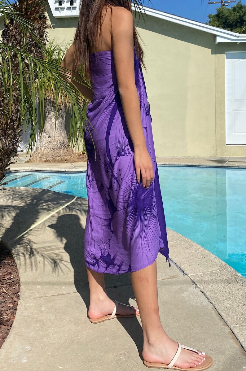 Op de afbeelding: Een paarse sarong met een bloemenprint. De sarong is om de taille gebonden en valt tot aan de enkels. De persoon draagt witte sandalen.
