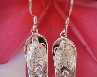 May include: Silver-coloured flip-flop earrings. Each earring has a detailed flip-flop design with floral and leaf patterns. The earrings hang from a simple hook.