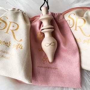 Op de afbeelding: Een lichtgekleurde houten pendel met een oneindigheidssymbool, hangend voor een roze trekkoordtas en twee off-white trekkoordtassen. De tassen hebben gouden tekst met de tekst "SR" en "Using your gifts". De website-adres is ook zichtbaar.