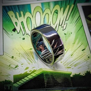 May include: A silver ring featuring a comic book character design. The ring has a textured, brushed finish and a black and green image of a superhero.