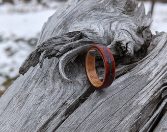 May include: A wooden ring with a red and blue inlay. The ring is sitting on a piece of driftwood.