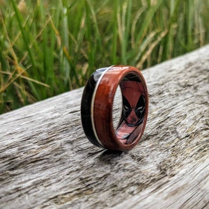 May include: A wooden ring with a black and red Deadpool design. The ring has a red and black wood grain pattern with a white stripe.