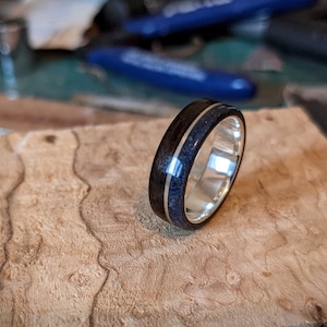 May include: A dark wood ring with a silver band and a blue inlay. The ring is sitting on a piece of wood with a unique grain pattern.