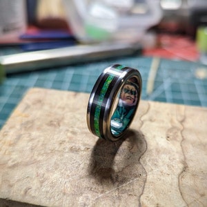 Può includere: Un anello nero e verde con una fascia argentata. L'anello presenta un design di personaggio dei fumetti all'interno.