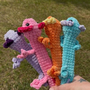 Puede incluir: Cuatro juguetes de peluche con forma de ajolote hechos de hilo suave y texturizado en tonos morado claro, rosa, naranja y azul claro. Cada criatura tiene ojos oscuros pequeños, branquias externas a los lados de la cabeza y cuatro extremidades.