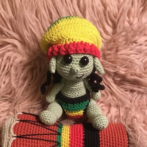 Puede incluir: Una muñeca elfo de crochet verde que lleva un gorro de crochet amarillo, rojo y verde. La muñeca está sentada en un tambor de crochet rojo, amarillo, verde y negro.