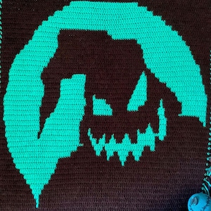 May include: Crocheted square with a teal green silhouette of Jack Skellington's face from the Nightmare Before Christmas movie. The background is brown.