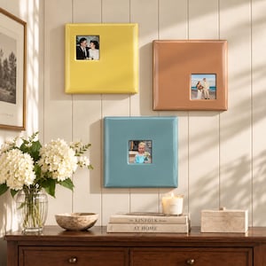 May include: Three square wall-mounted photo frames in yellow, copper, and blue, each featuring a centered or offset square cutout for a photograph. The frames have a smooth, glossy finish and are arranged vertically on a paneled wall surface.