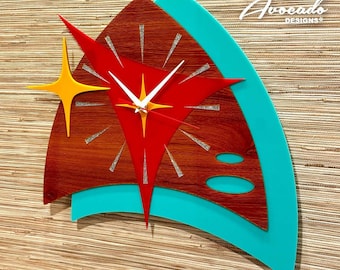 Puede incluir: Un reloj de madera, rojo y turquesa con un diseño de estrella y detalles de purpurina plateada. La esfera del reloj tiene una aguja horaria blanca, una aguja minutera amarilla y una estrella amarilla en el centro. El reloj está etiquetado como "Atomic Avocado Designs".