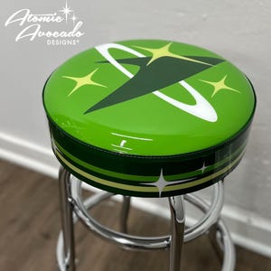 May include: A green bar stool with a chrome base. The seat is upholstered with a green vinyl fabric and features a retro space-age design with a white and green planet and yellow stars. The stool is labeled "Atomic Avocado Designs".