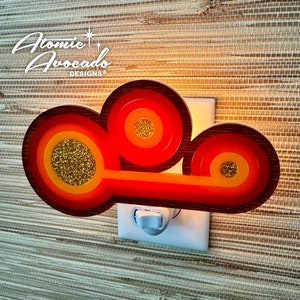 May include: A retro-style night light shaped like a cloud with three concentric circles in shades of orange, red, and yellow. The circles have a glittery gold center. The night light is plugged into a white electrical outlet.