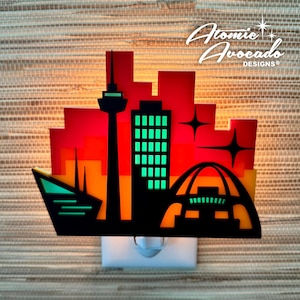 May include: A night light featuring a colorful cityscape silhouette. The design includes a tower, buildings with green windows, and an arched structure, set against a gradient of red, orange, and yellow. The night light has a white base and the text "Atomic Avocado Designs".