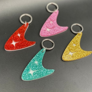Könnte beinhalten: Vier Schlüsselanhänger in Form eines Bumerangs mit Glitzer in verschiedenen Farben: Rot, Pink, Gelb und Türkis.
