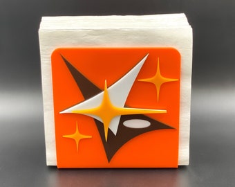 May include: Orange and white geometric design napkin holder with a starburst pattern. The holder is made of plastic and has a modern design.