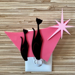 May include: A pink and black nightlight with a starburst design and three black cat silhouettes. The nightlight is plugged into an electrical outlet.