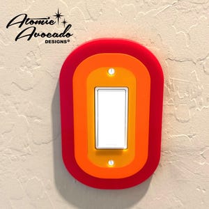 May include: A red, orange, and yellow light switch cover with a white light switch in the center. The cover has a retro, 70s style design.