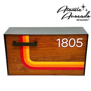 Puede incluir: Un buzón de madera con un diseño retro que presenta una franja naranja, amarilla y roja curvada. El buzón tiene una solapa de metal negro y un número blanco "1805" en la parte delantera. El buzón está hecho por Atomic Avocado Designs.