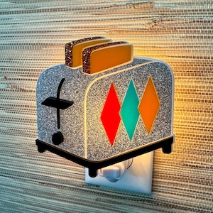Op de afbeelding: Een zilveren glitter-toaster nachtlampje met een stekkerbasis. De toaster heeft drie sneetjes brood in de gleuven en een rood, turquoise en oranje ruitpatroon aan de voorkant.