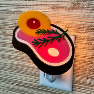 May include: A pink and black nightlight shaped like a steak with a yellow pineapple on top. The nightlight is plugged into an outlet.