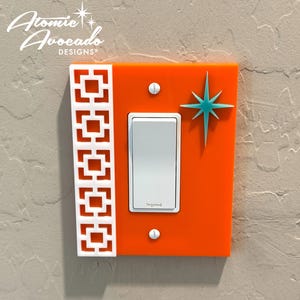 Puede incluir: Tapa de interruptor de luz naranja con un diseño geométrico blanco en el lado izquierdo y una estrella turquesa en el lado derecho. El interruptor es blanco y dice "Legrand".