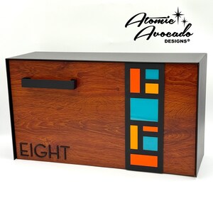 May include: A wooden mailbox with a dark finish and a black handle. The front features a colourful geometric design with orange, teal, and yellow shapes. The word "EIGHT" is cut out of the wood. The top right corner has the text "Atomic Avocado DESIGNS®".